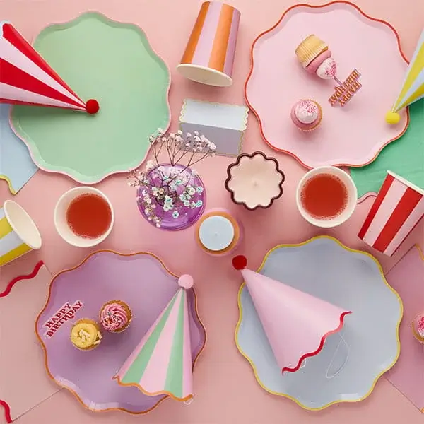 Pastel birthday party tableware with colorful plates, cups, napkins, and gold Happy Birthday lettering on a soft pink background